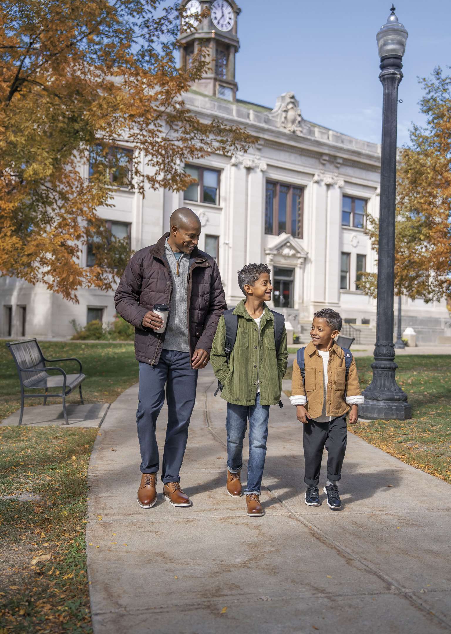 The featured image is a man wearing Nunn Bush shoes while walking on a paved path in front of a white building with two young boys in warm jackets.